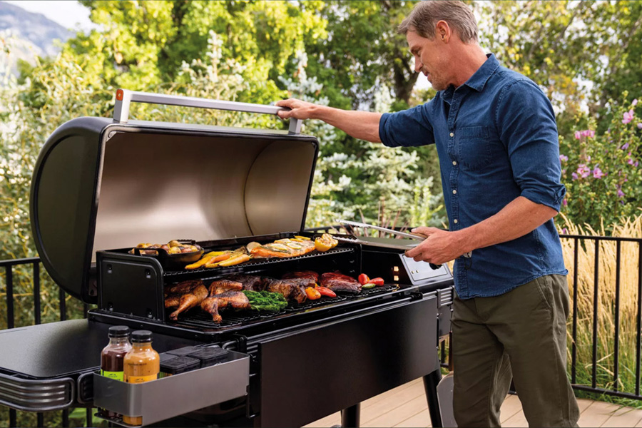 Auszeit - Wellness - Angebote - Grills. Mann am Traeger Grill mit diversen Grillsachen auf dem Rost